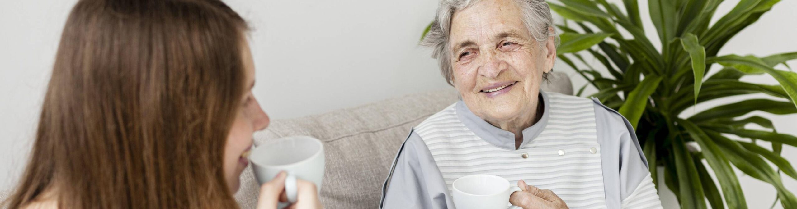 Elderly lady next to a plant, looking and smiling at her carer with a hot drink in a mug.