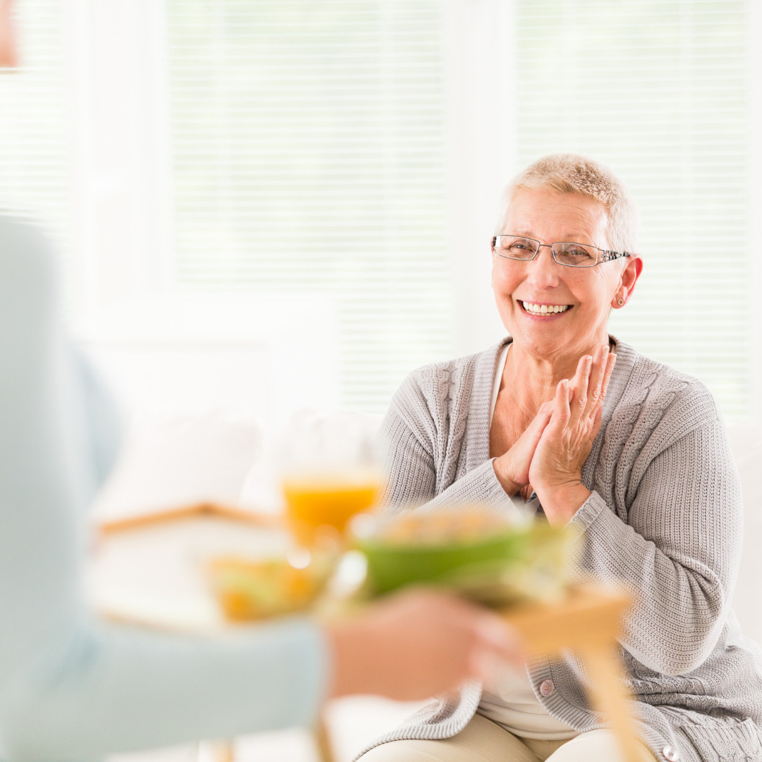 Elderly lady looking happy with hands together while carer carries in a tray with food and drink on.