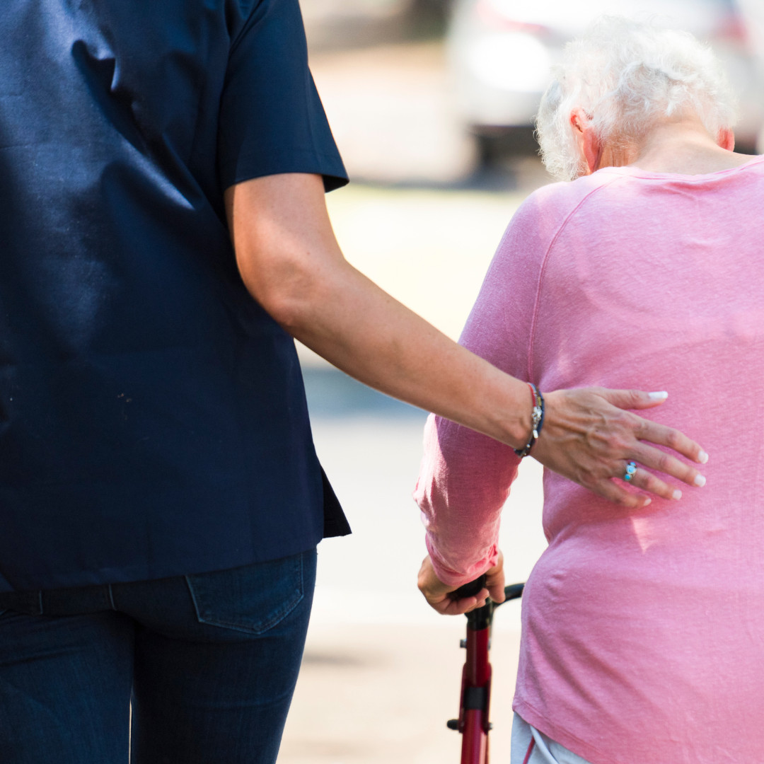 Backshot image of carer holding elderly ladies back while walking outside with a walking frame